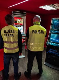 Zdjęcie kolorowe: policjant i funkcjonariusz KAS zabezpieczają nielegalne automaty do gier.
