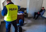 Policjant z zatrzymanym mężczyzna, który siedzi na krześle  w rogu pomieszczenia